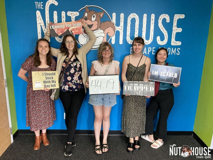 Five women hold signs in front of the Nuthouse Escape Rooms logo celebrating their 44:19 time
