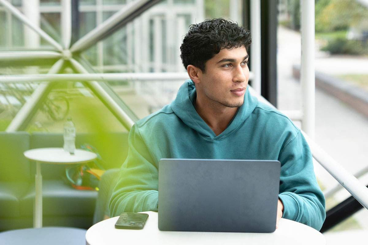 A man sitting at a table with a laptop, studying at the University of Rochester.