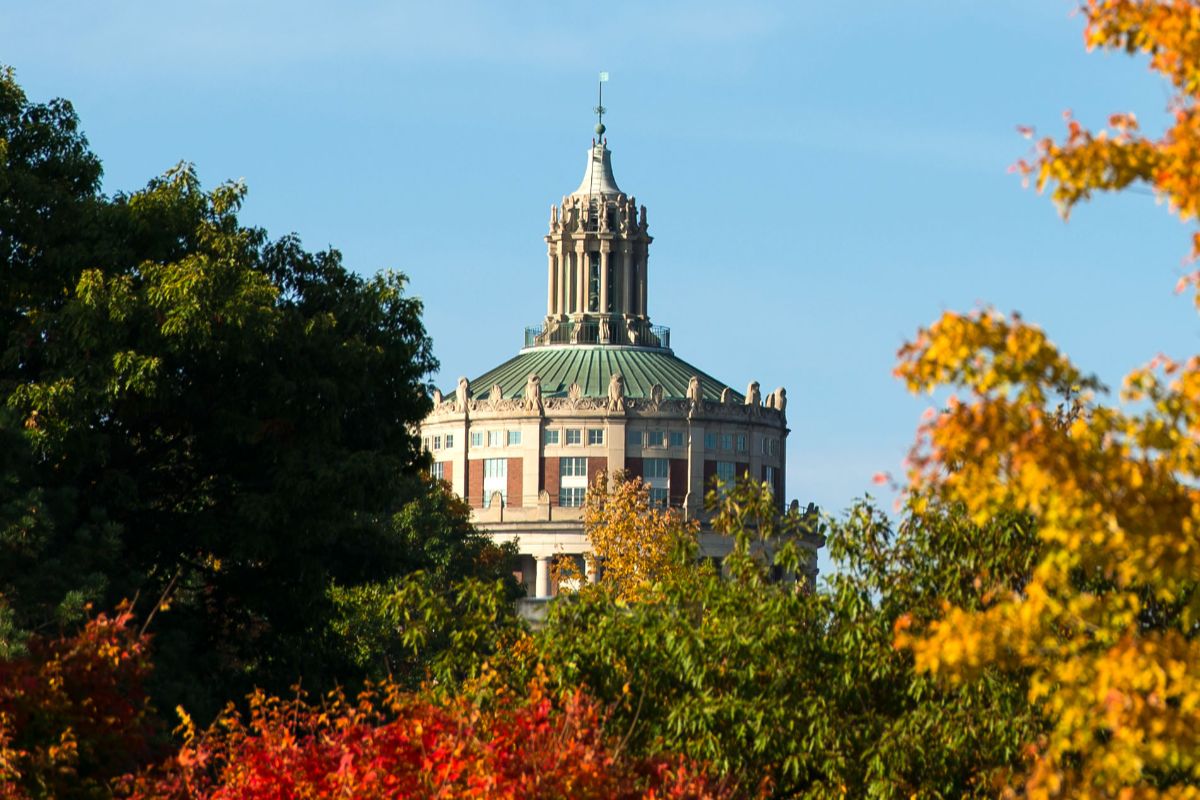 The University of Rochester library building surrounded by vibrant fall foliage, showcasing autumn colors and clear blue skies.