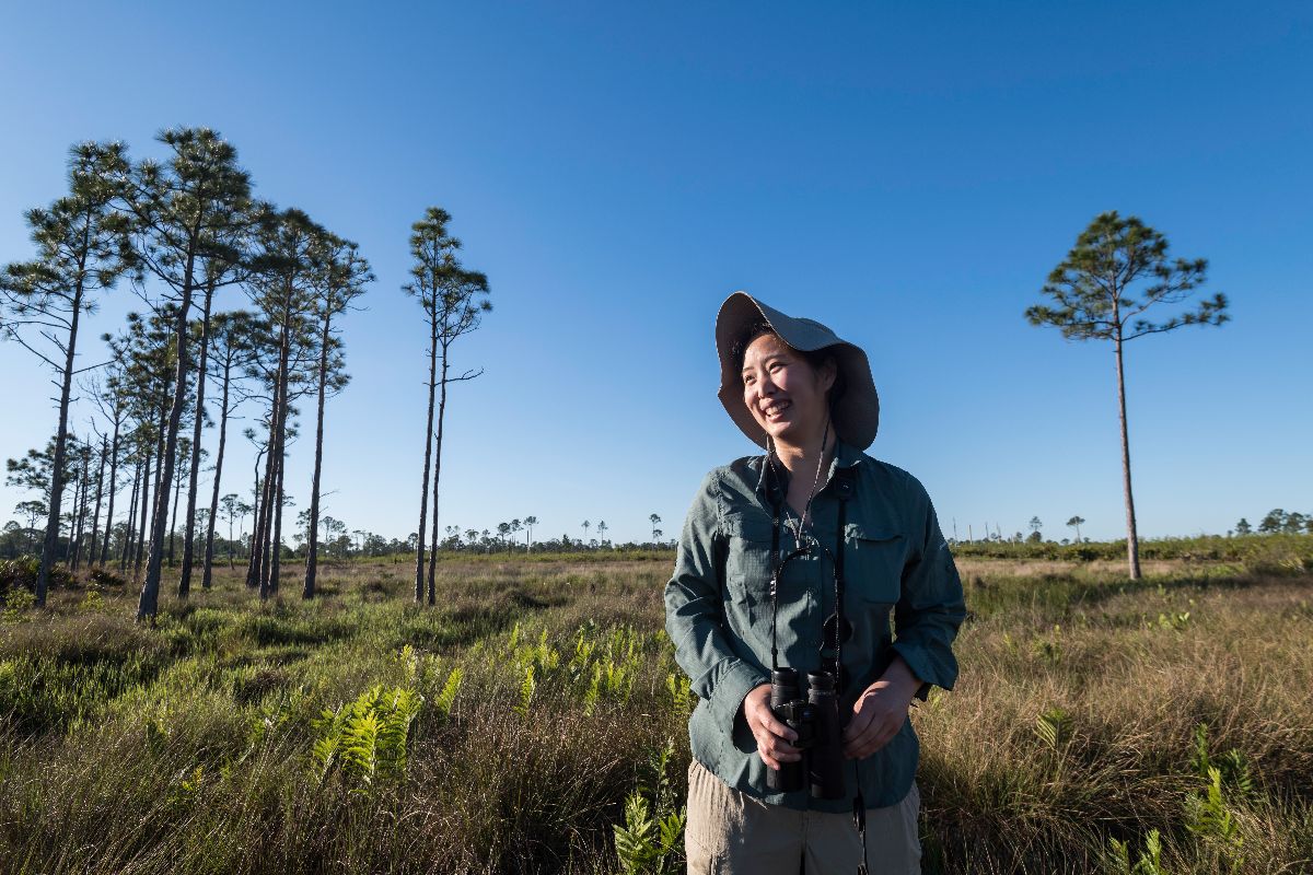 A woman wearing a hat stands in a grassy field surrounded by trees, enjoying the outdoors.