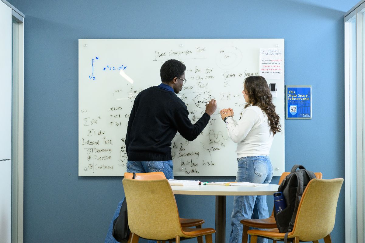 Two individuals collaboratively writing on a whiteboard in a university classroom setting.